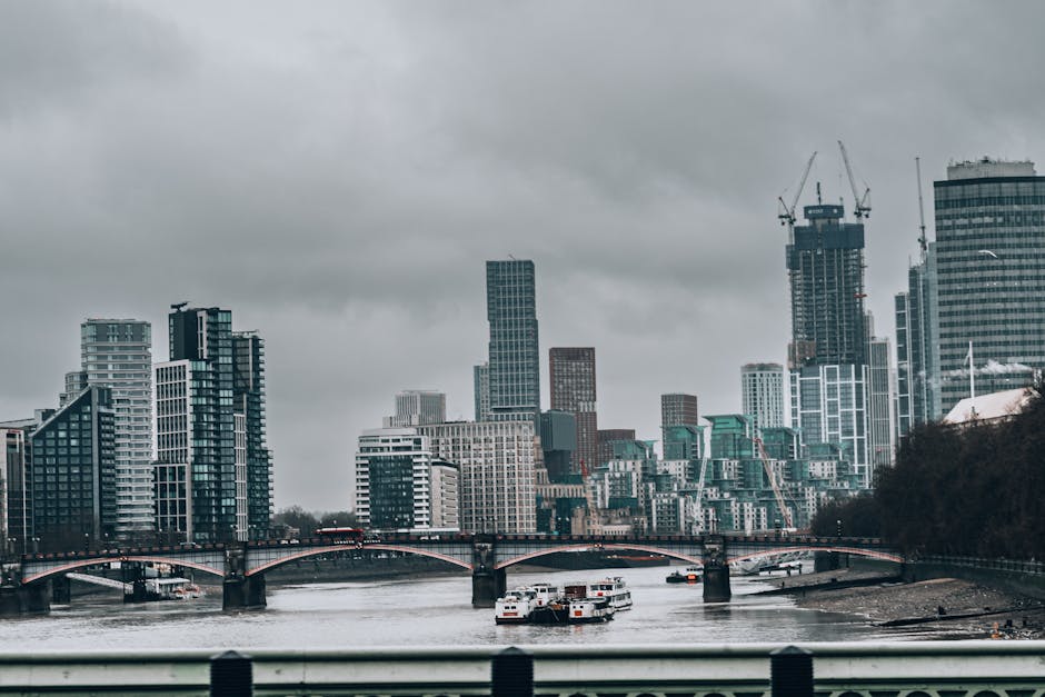 A wide view of the London skyline along the River Thames during daylight, featuring modern high-rise buildings with a mix of glass and concrete facades. In the foreground, several boats are moored alo